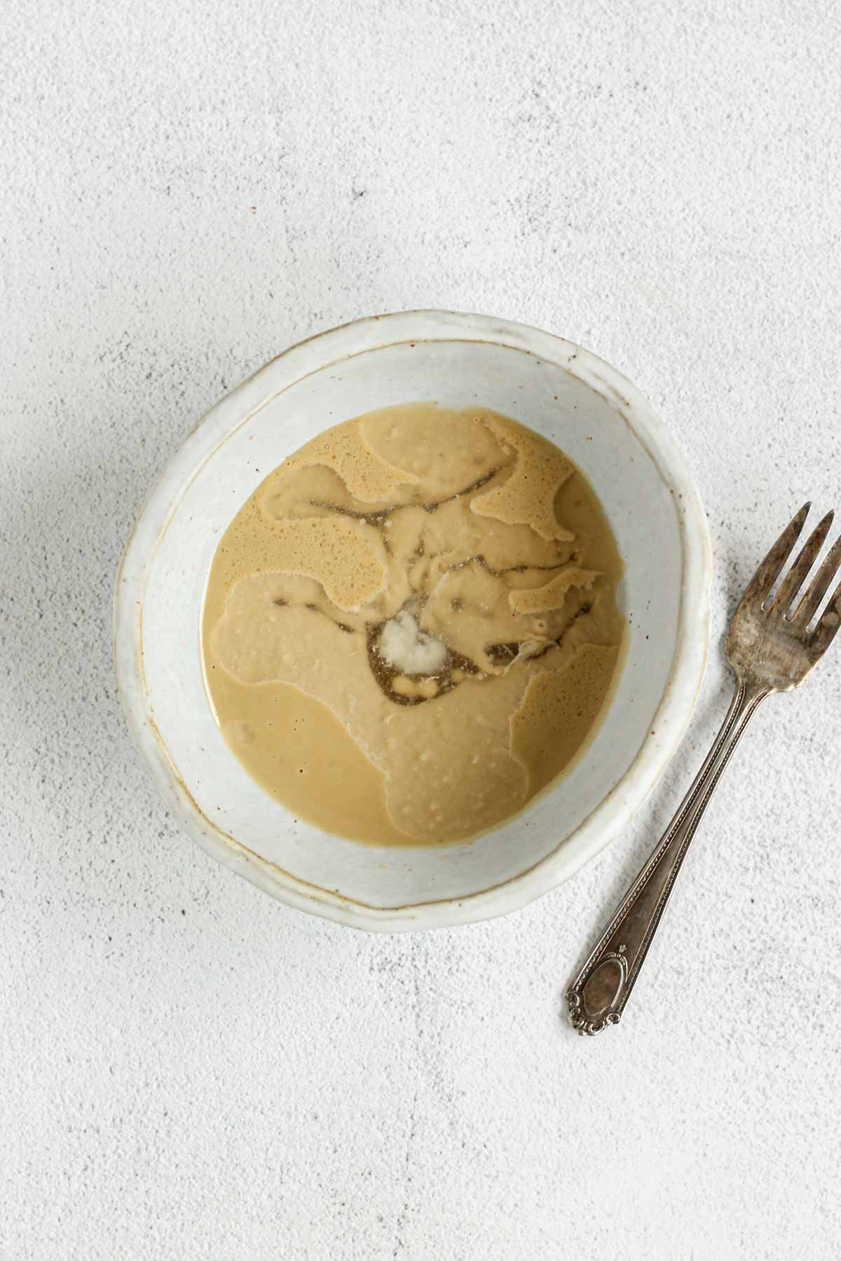 Maple tahini salad dressing ingredients in a shallow bowl unmixed with a fork on the side of the bowl.
