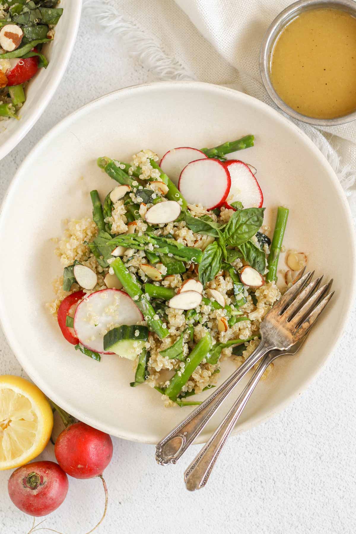 Quinoa and asparagus salad with two forks on a small plate with a side of dressing and half a lemon and radishes off to the side.