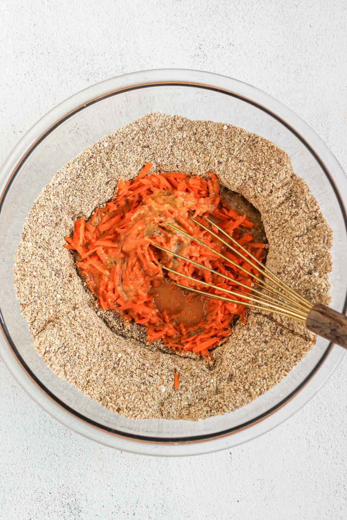 Glass mixing bowl with dry ingredients for carrot flaxseed muffins with wet ingredients in the center being mixed with a wire whisk.