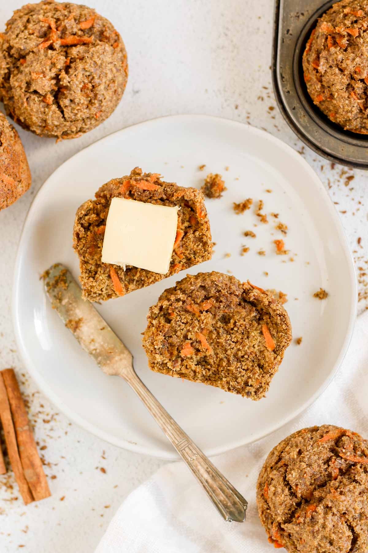 Round plate with a carrot flaxseed muffin cut in half with a square of butter on one half and a small knife resting on the plate with additional muffins on the side.
