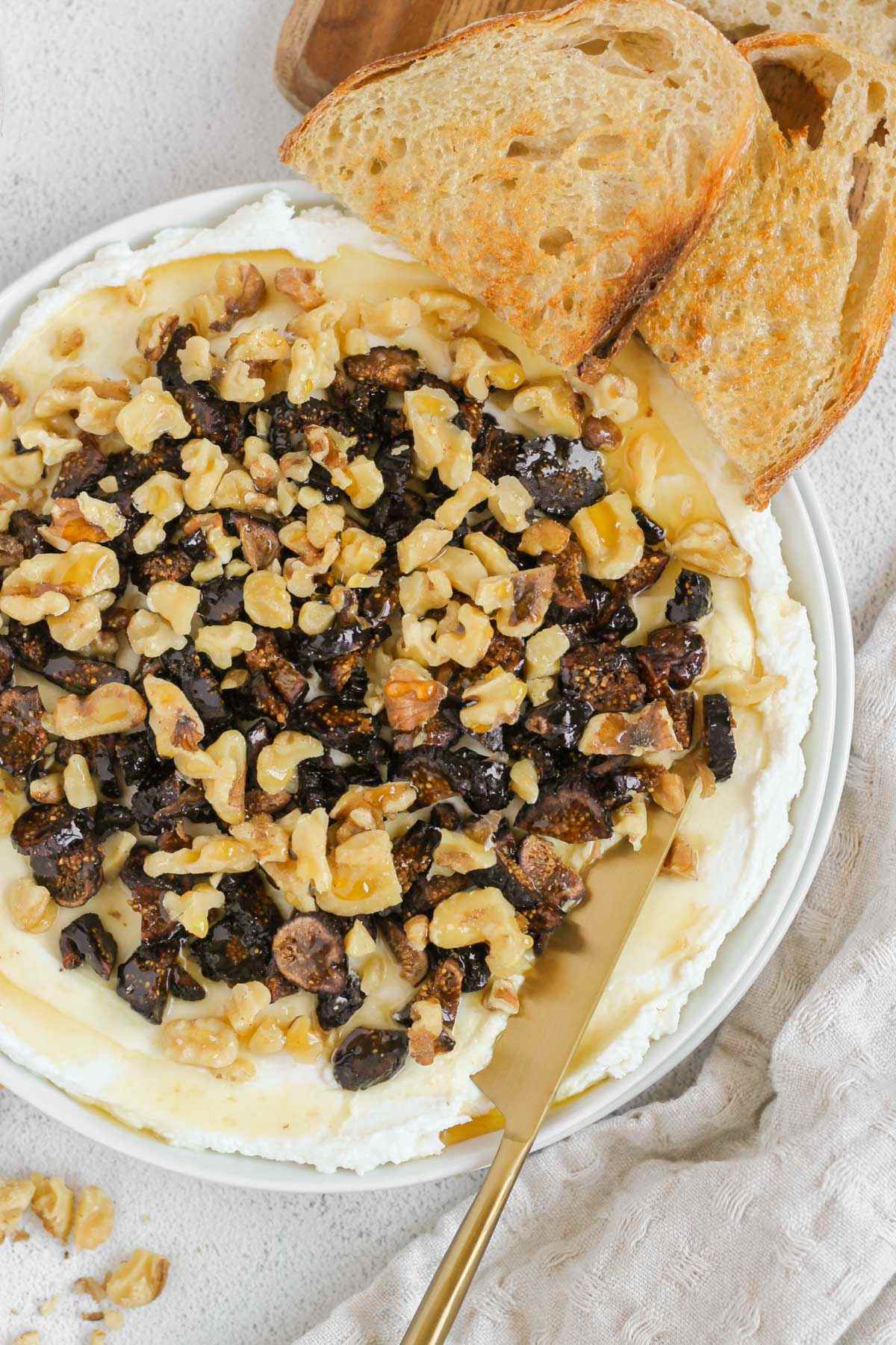 Whipped goat cheese topped with honey, sliced dried figs and walnuts on a small round plate. with a knife scooping up some of the spread and 2 pieces of toast on the side.