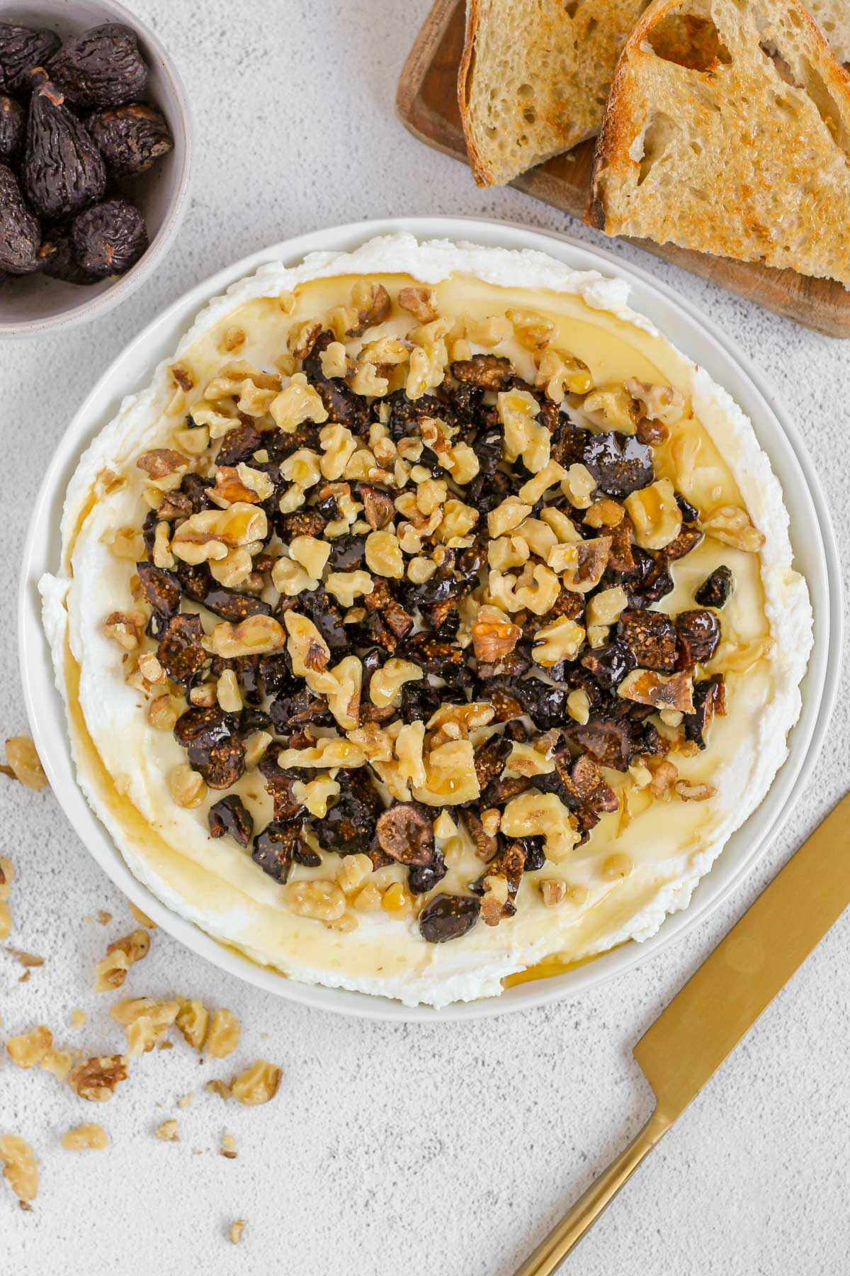 Whipped goat cheese topped with honey, sliced dried figs and walnuts on a small round plate with toast and a gold knife on the side.