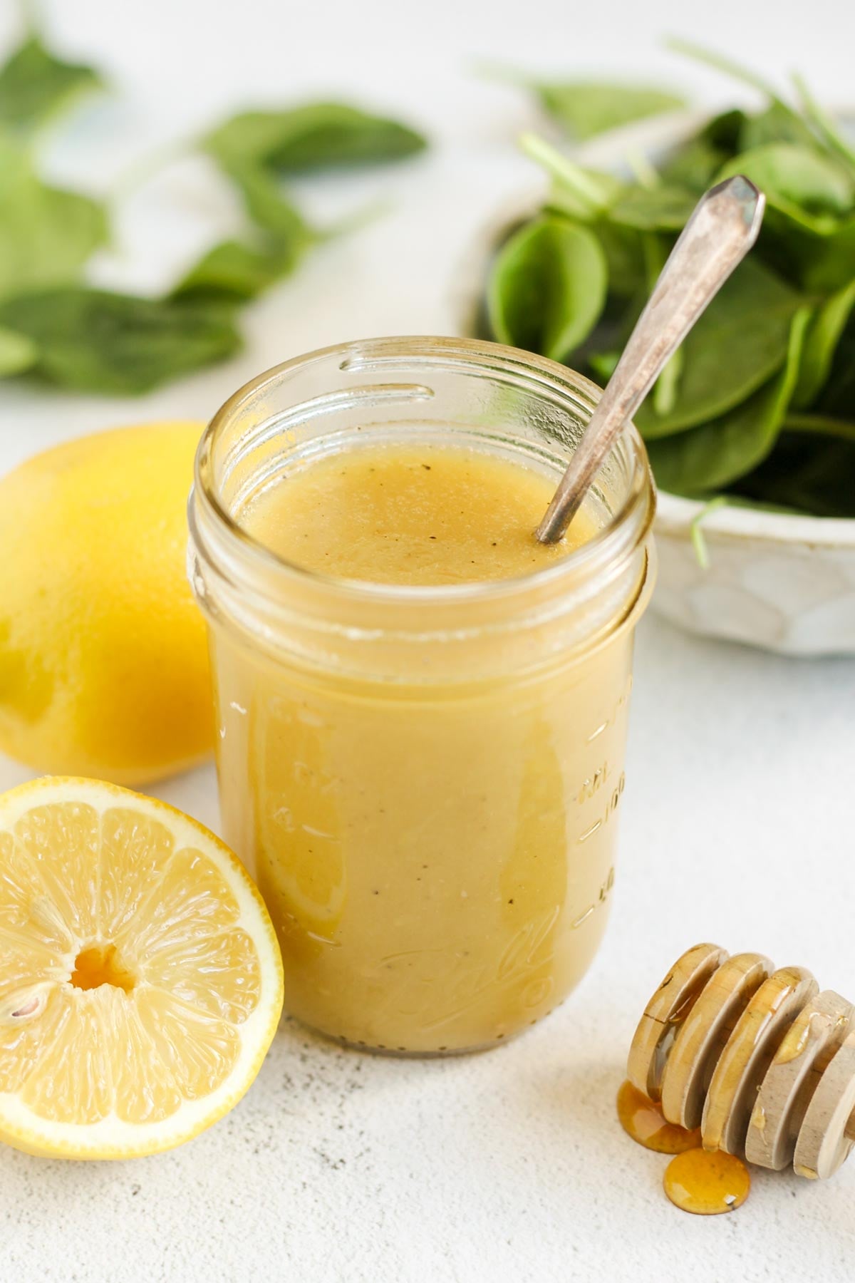 Lemon honey dressing in a mason jar with a sliced lemon, honey dipper and a bowl of spinach on the side.
