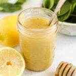 Lemon honey dressing in a mason jar with a sliced lemon, honey dipper and a bowl of spinach on the side.