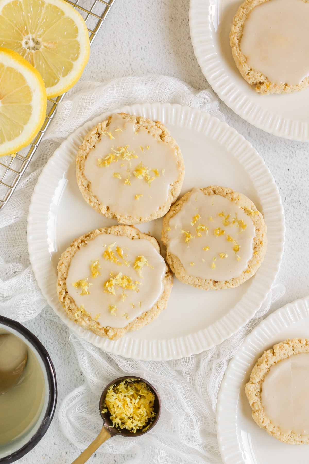 Three glazed lemon cookies sprinkled with lemon zest on a small plate with additional plates, lemons, and a bowl of glaze off to the sides.