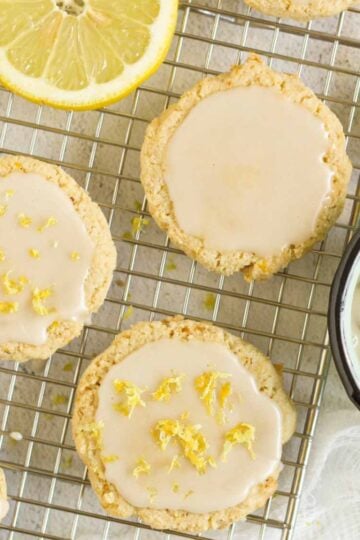 Almond flour lemon cookies topped with a glaze on a rectangular wire cooling rack with slices of lemon placed on the rack.
