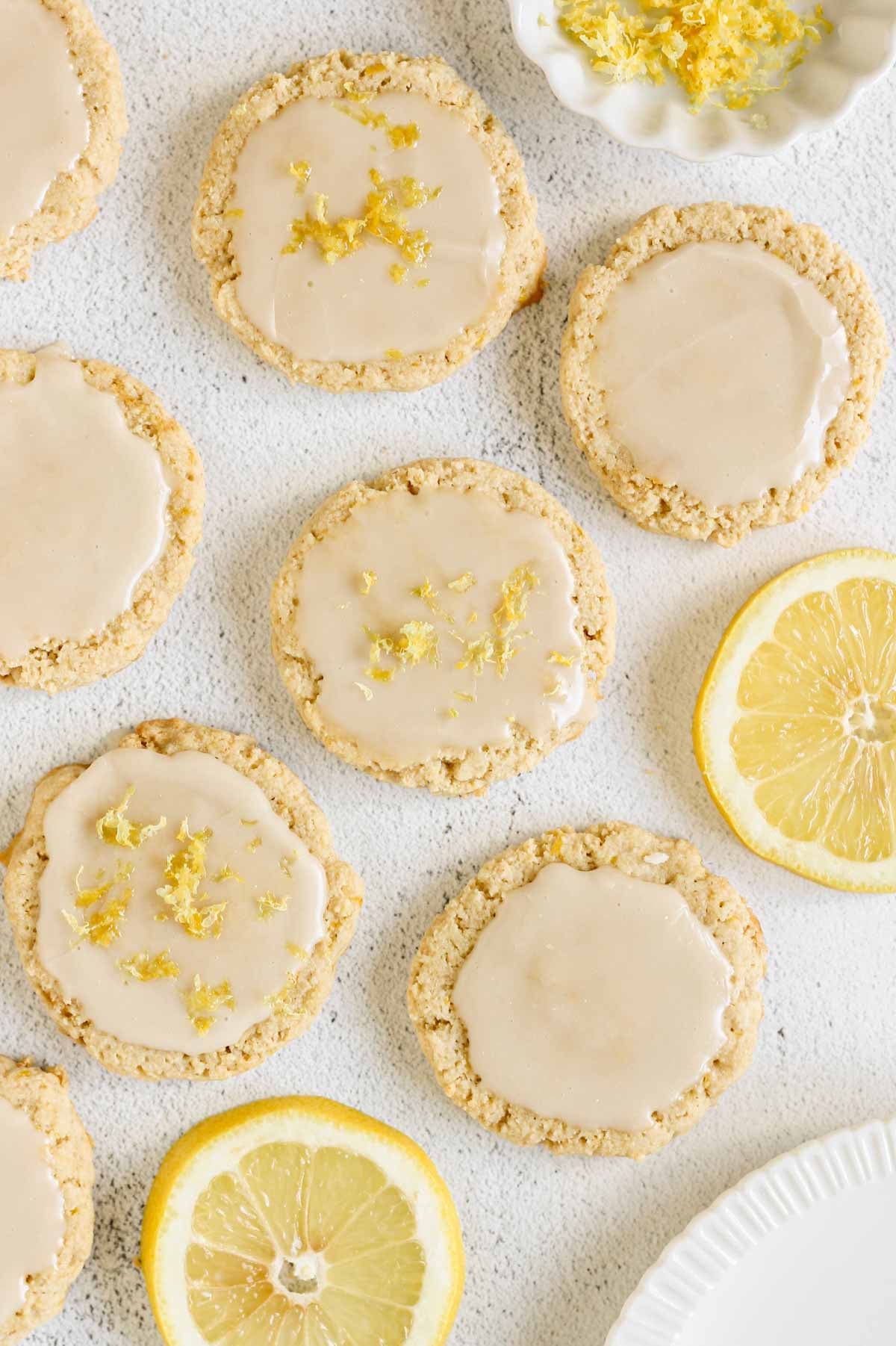 Glazed almond flour lemon cookies topped with fresh lemon zest, arranged on a light surface with sliced lemons and a small bowl of lemon zest nearby.
