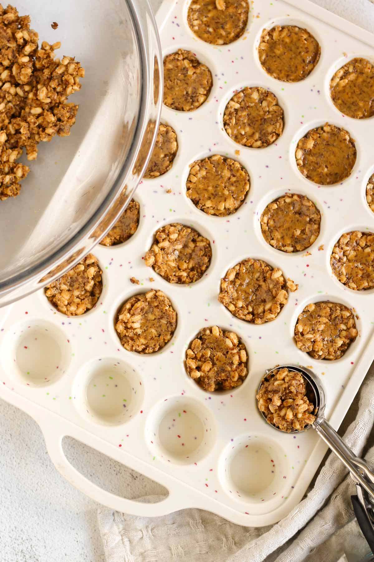 24-cup silicone muffin pan partially filled peanut butter brown rice crisp mix with a spring loaded scoop scooping up some mix.