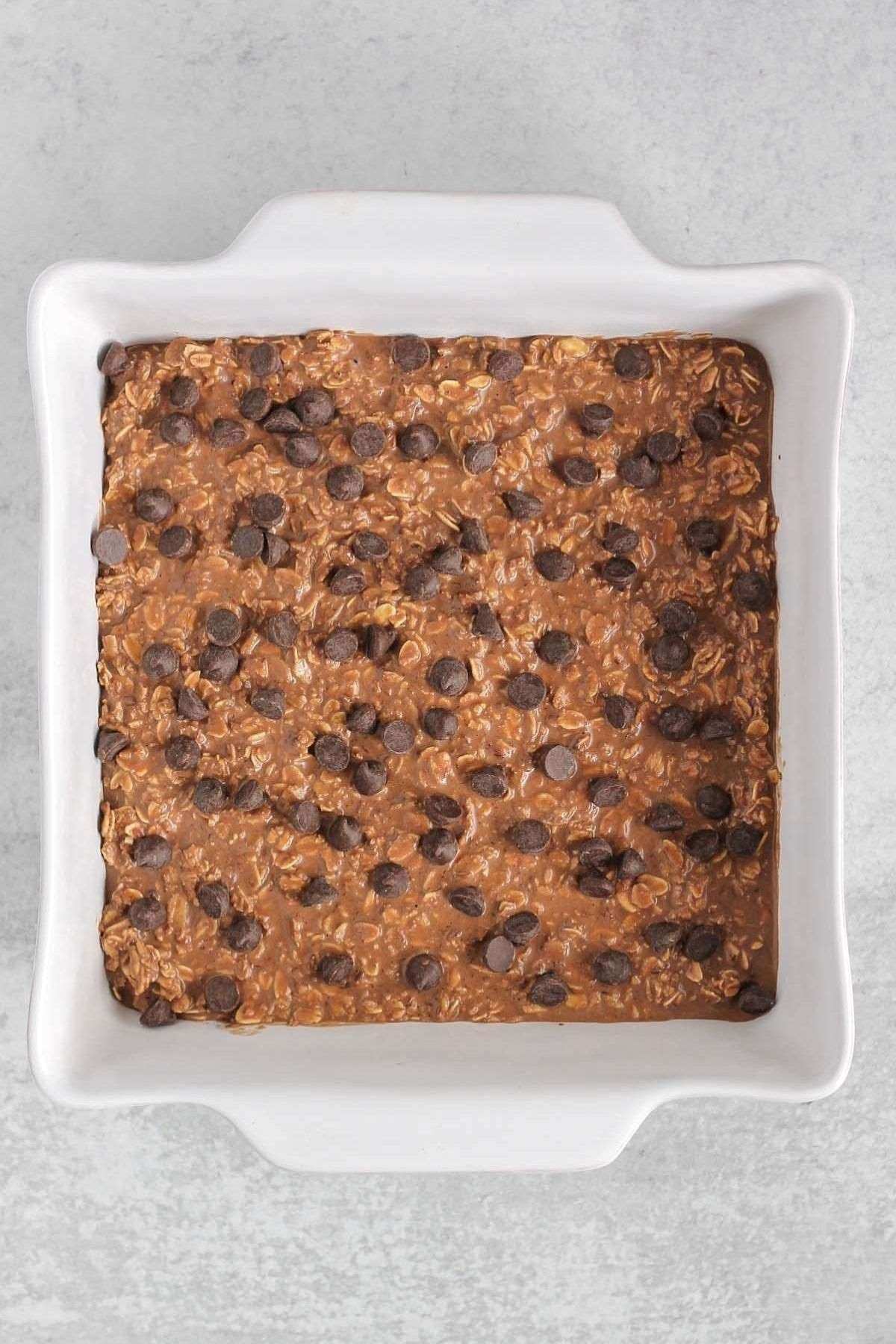 Chocolate baked oats mixture topped with chocolate chips, before being baked, in a square baking dish.