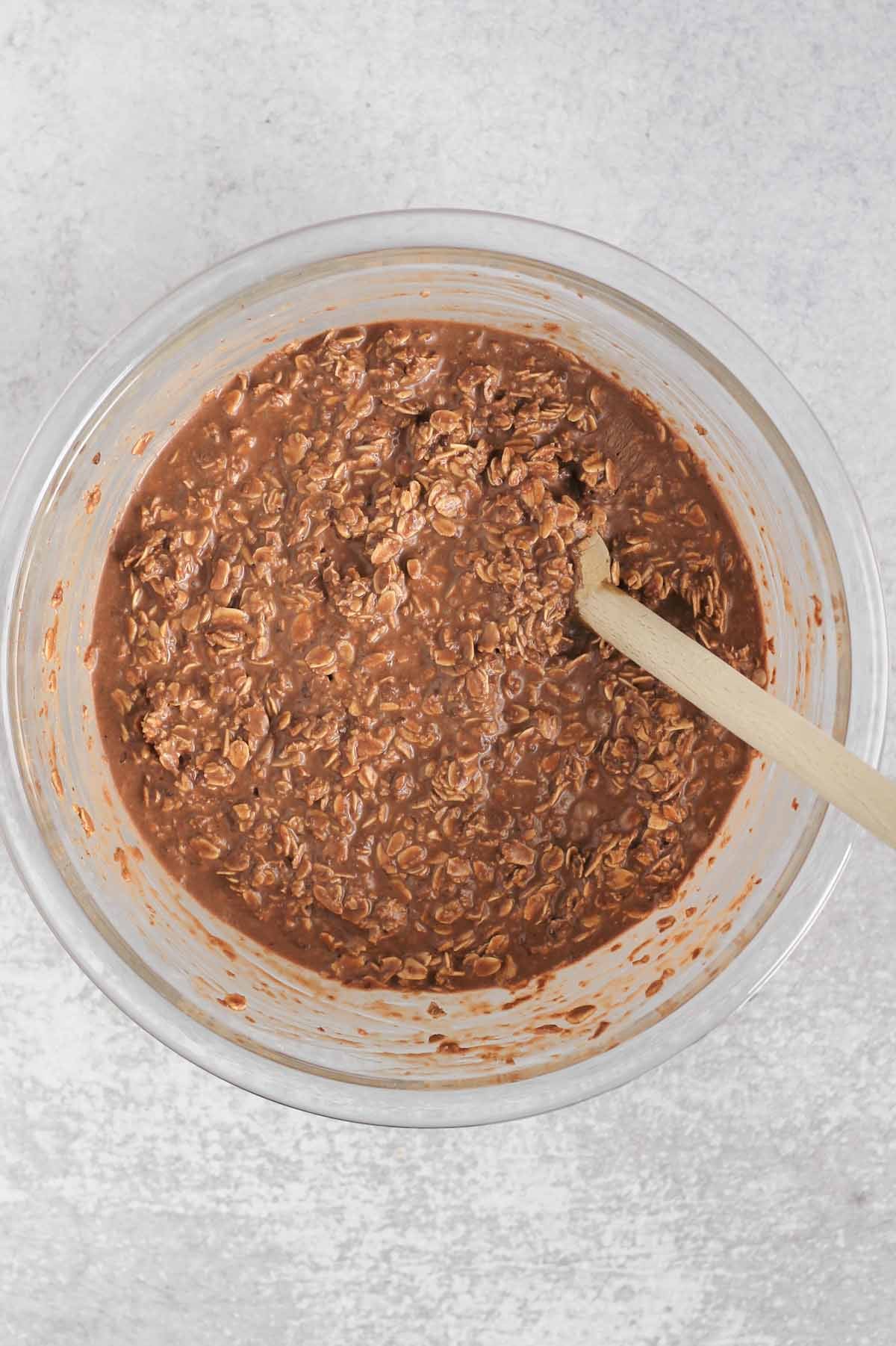 Chocolate baked oats ingredients mixed together in a glass mixing bowl with a wooden spoon.