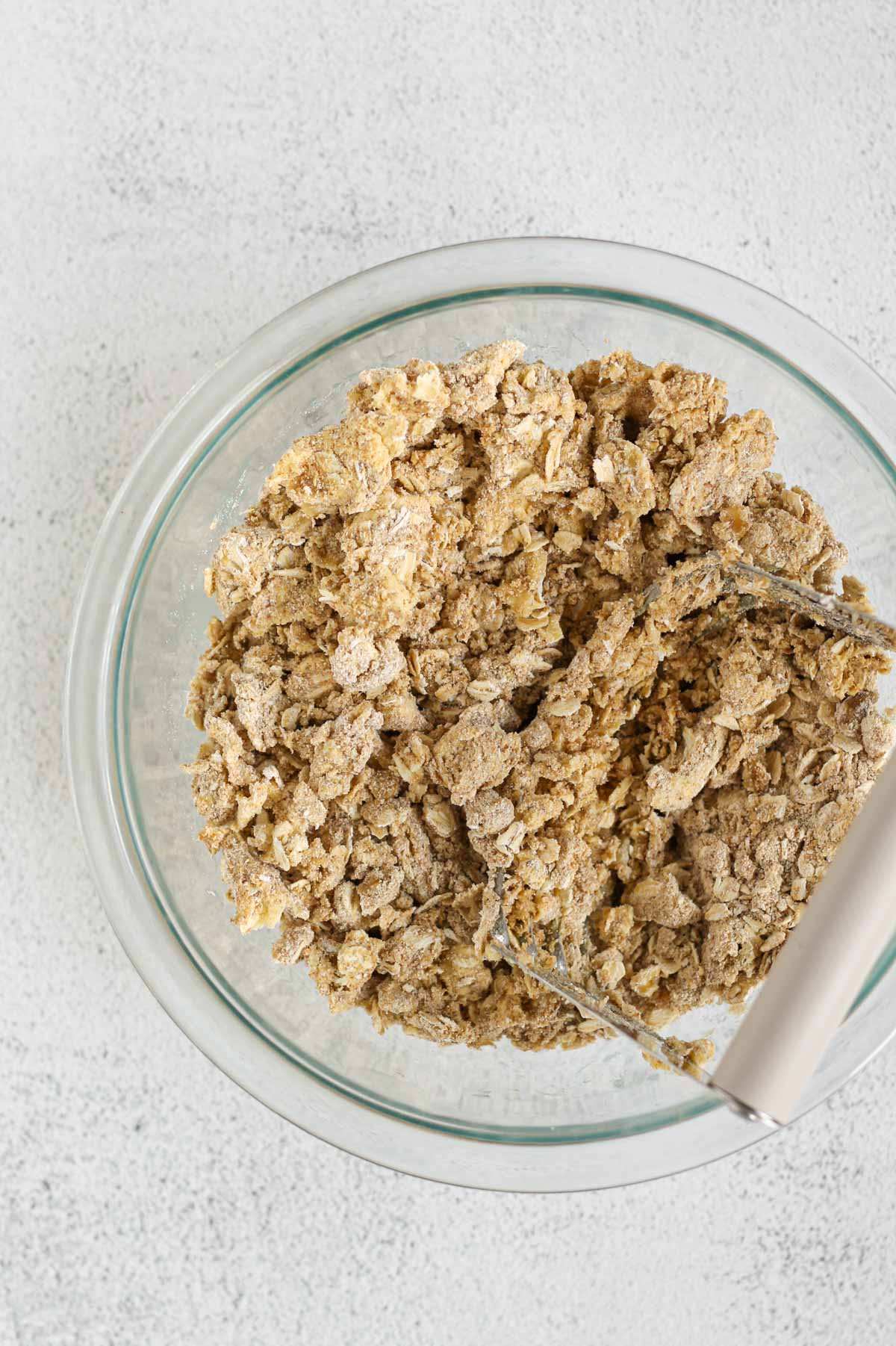 Pastry cutter cutting butter into a mixture of oats, oat flour and coconut sugar in a small glass mixing bowl.