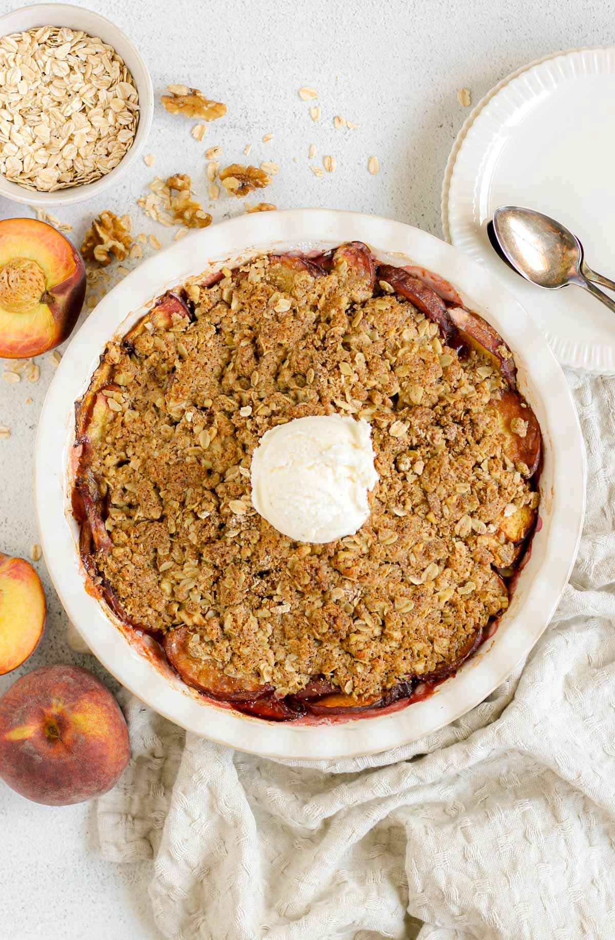 Baked peach crisp topped with a scoop of ice cream in a ceramic pie dish with sliced peaches, plates, and a bowl of oats scattered on the sides.