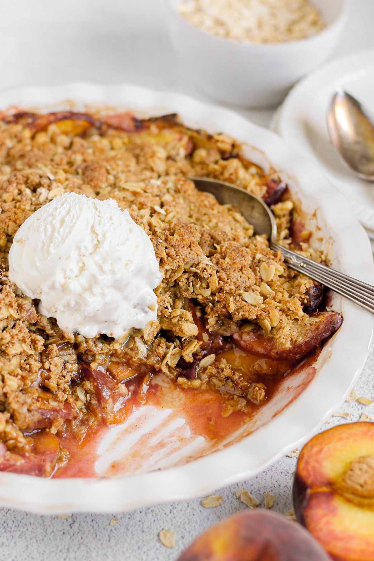 A baked peach crisp in a white baking dish topped with a scoop of vanilla ice cream. A serving has been scooped out, revealing juicy peaches beneath a golden oat crumble topping. Fresh peach halves and scattered oats sit nearby.