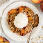 Serving of peach crisp on a round plate topped with ice cream and a vintage spoon on the plate with sliced peaches on the sides of the plate.
