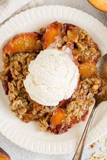 Serving of peach crisp on a round plate topped with ice cream and a vintage spoon on the plate with sliced peaches on the sides of the plate.