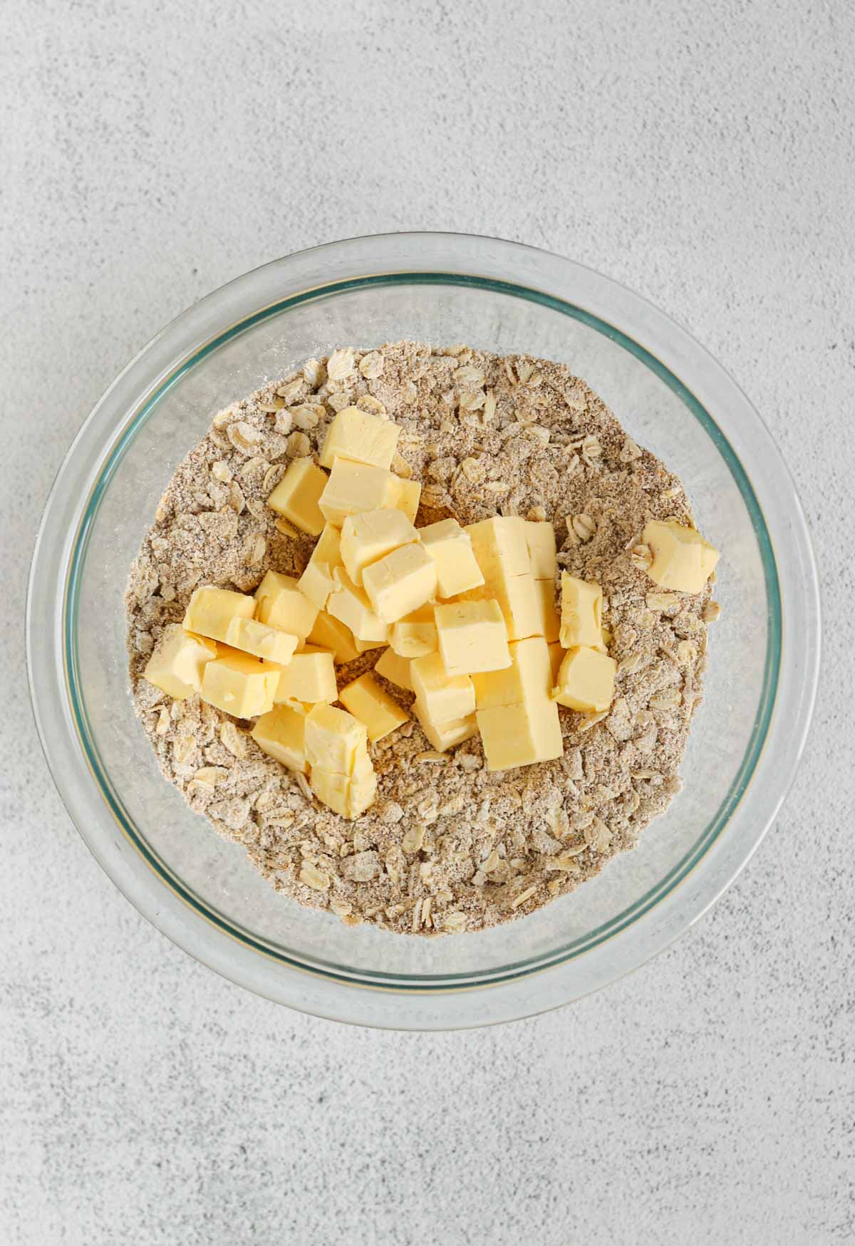Oats, oat flour, and coconut sugar mixed in a small glass mixing bowl with cubes of butter added to the bowl.
