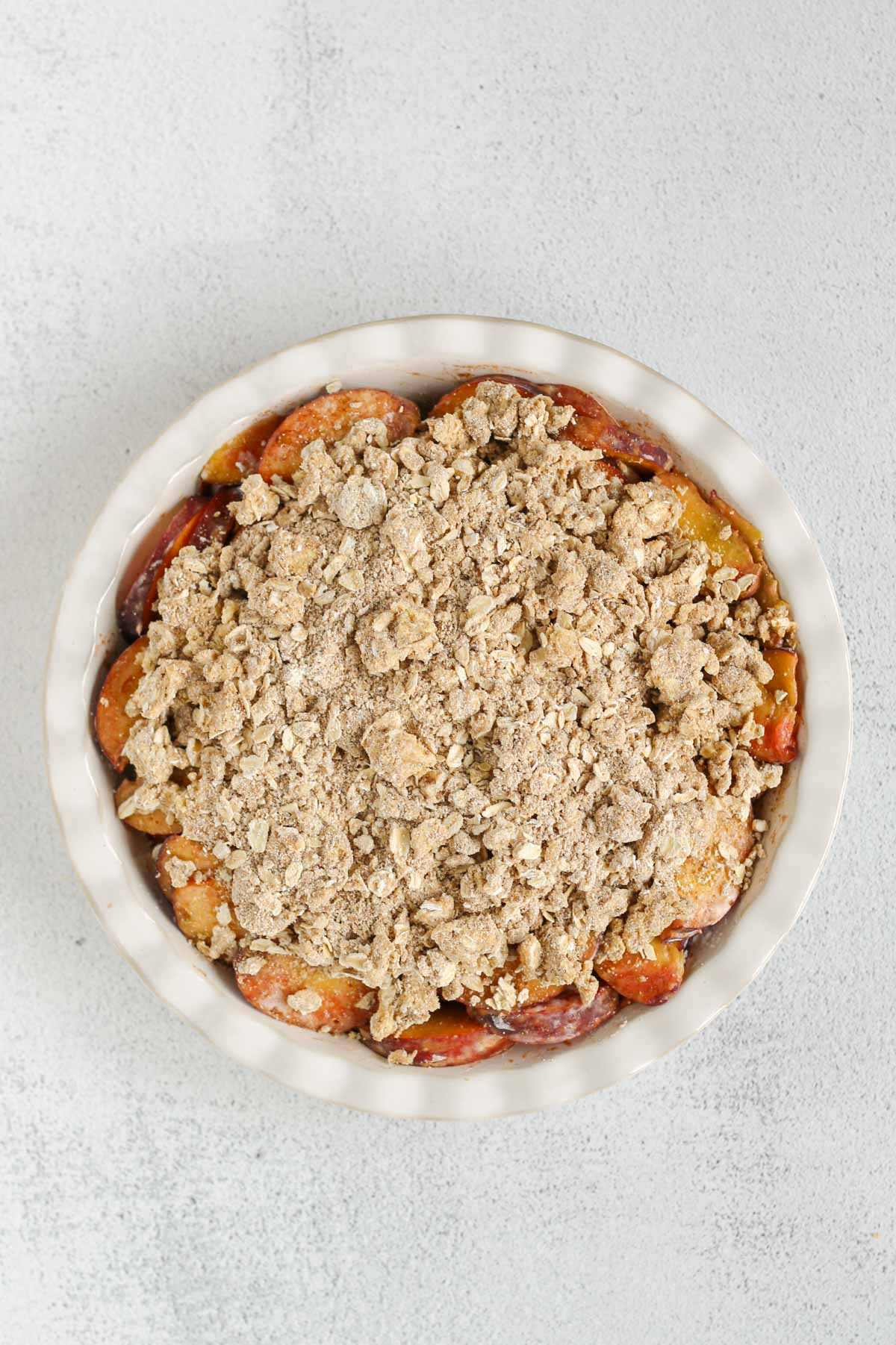 Sliced peaches in a ceramic pie dish topped with oat flour, oat, and butter mixture.