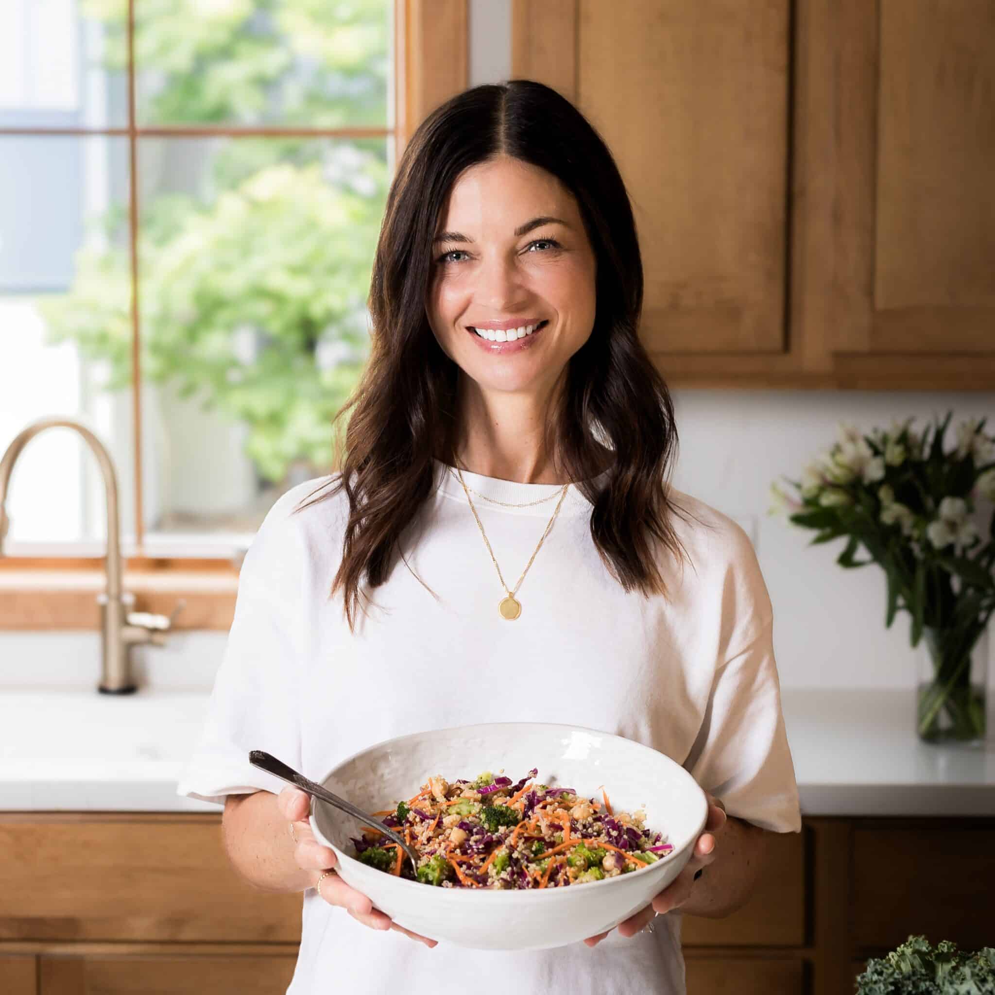 A woman with shoulder length brown hair holding a serving bowl full of colorful quinoa salad.
