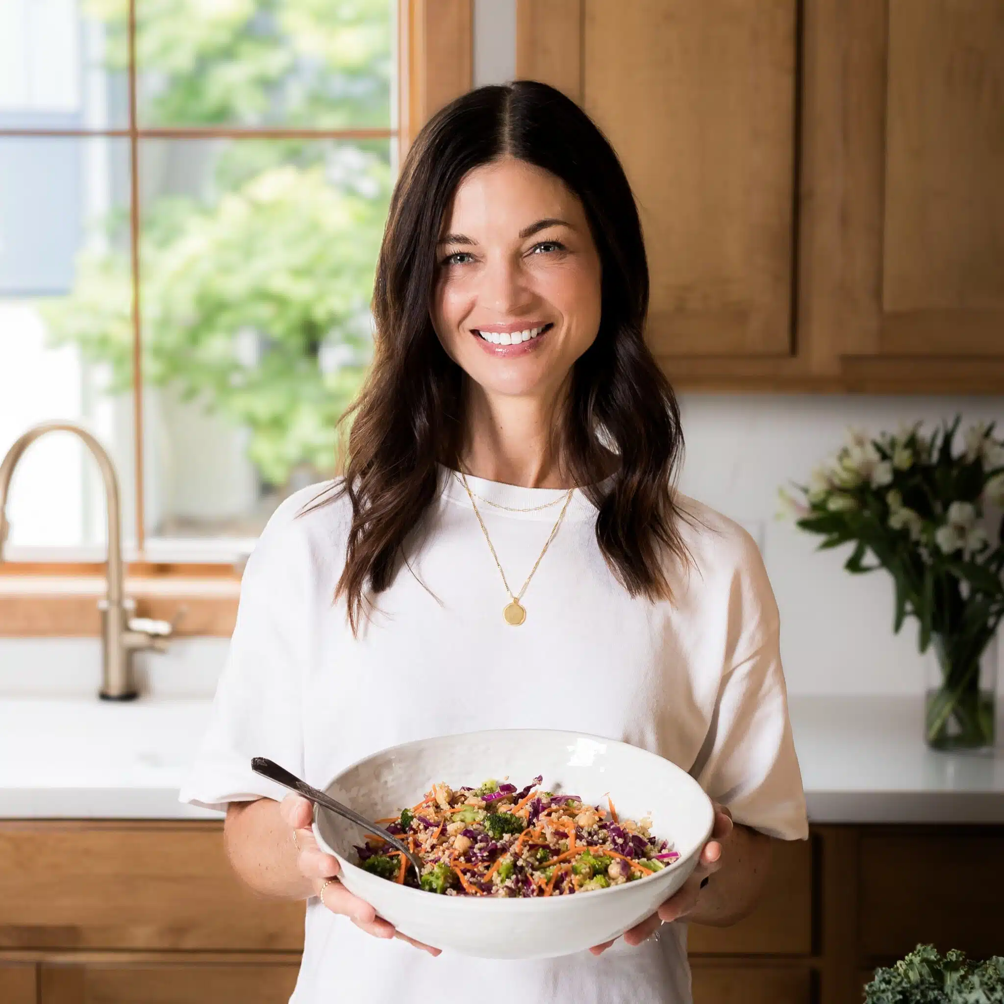A woman with shoulder length brown hair holding a serving bowl full of colorful quinoa salad.