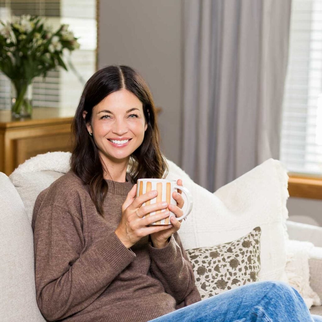 A woman with shoulder length brown hair holding sitting on a couch holding a coffee mug.