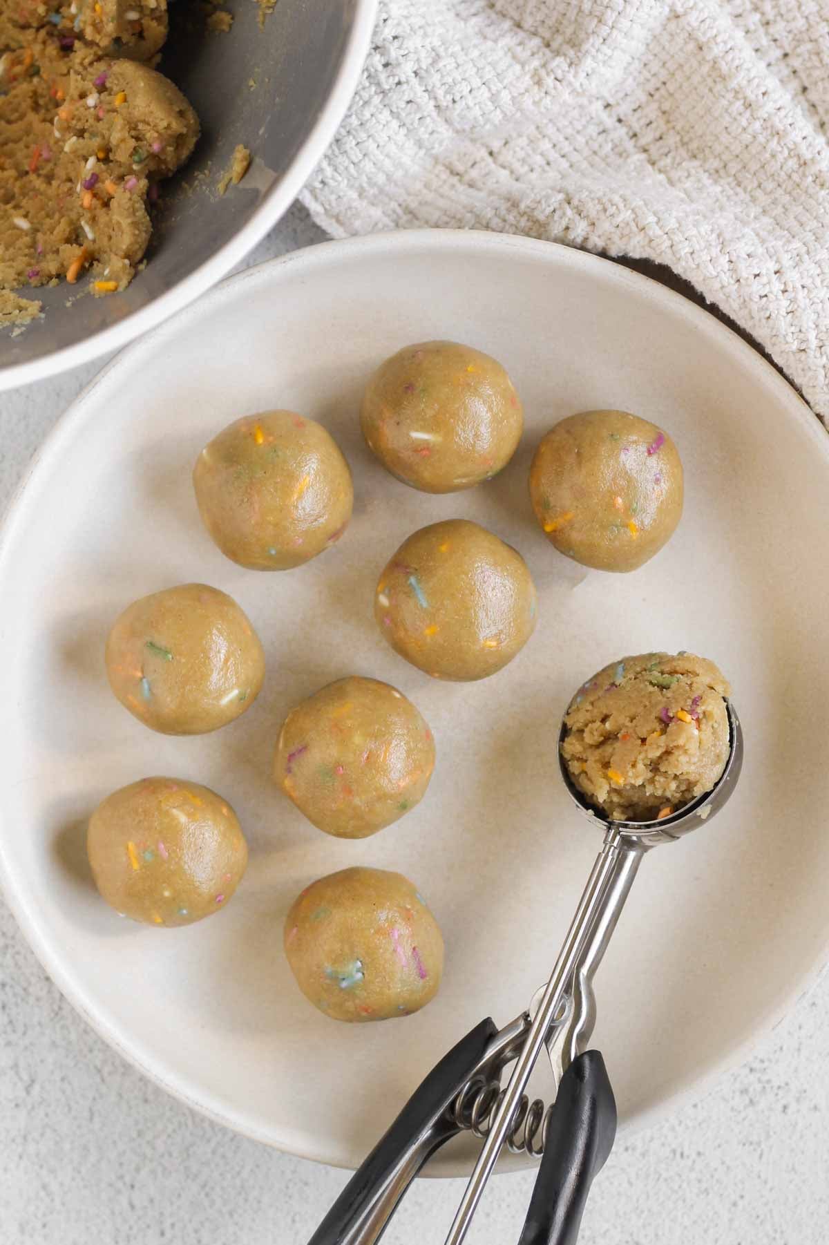 Birthday cake protein balls rolled on a large plate with a spring-loaded scoop filled with protein ball mix on the side of the plate.