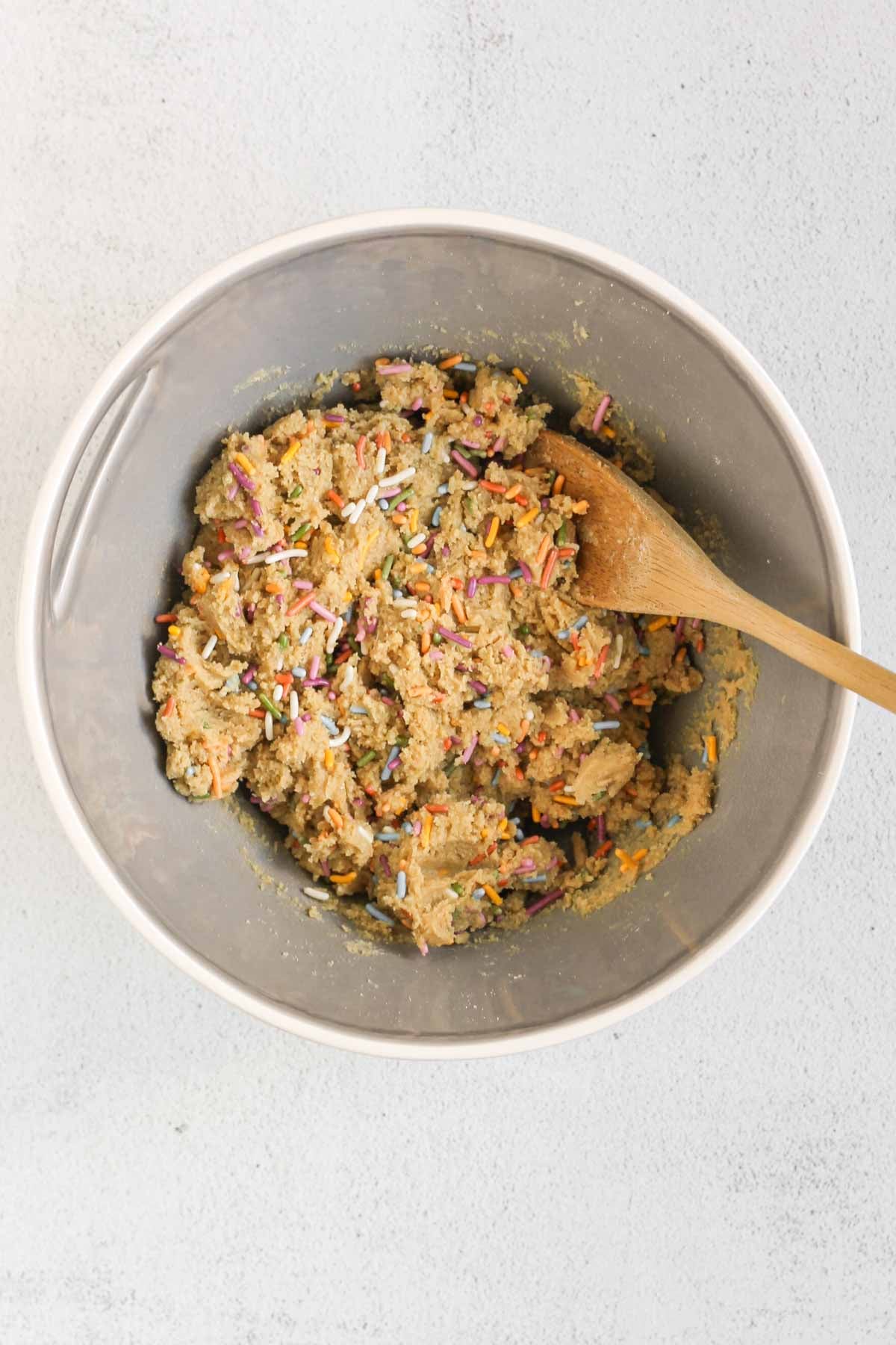 Birthday cake protein ball mix, with sprinkles, being mixed in a mixing bowl with a wooden spoon.