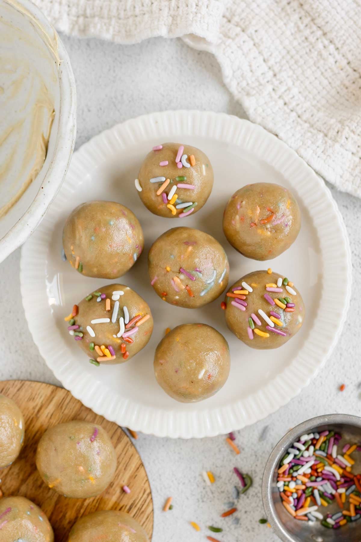 Six birthday cake protein balls on a small round plate with a small dish of sprinkles and a dish towel off to the side.