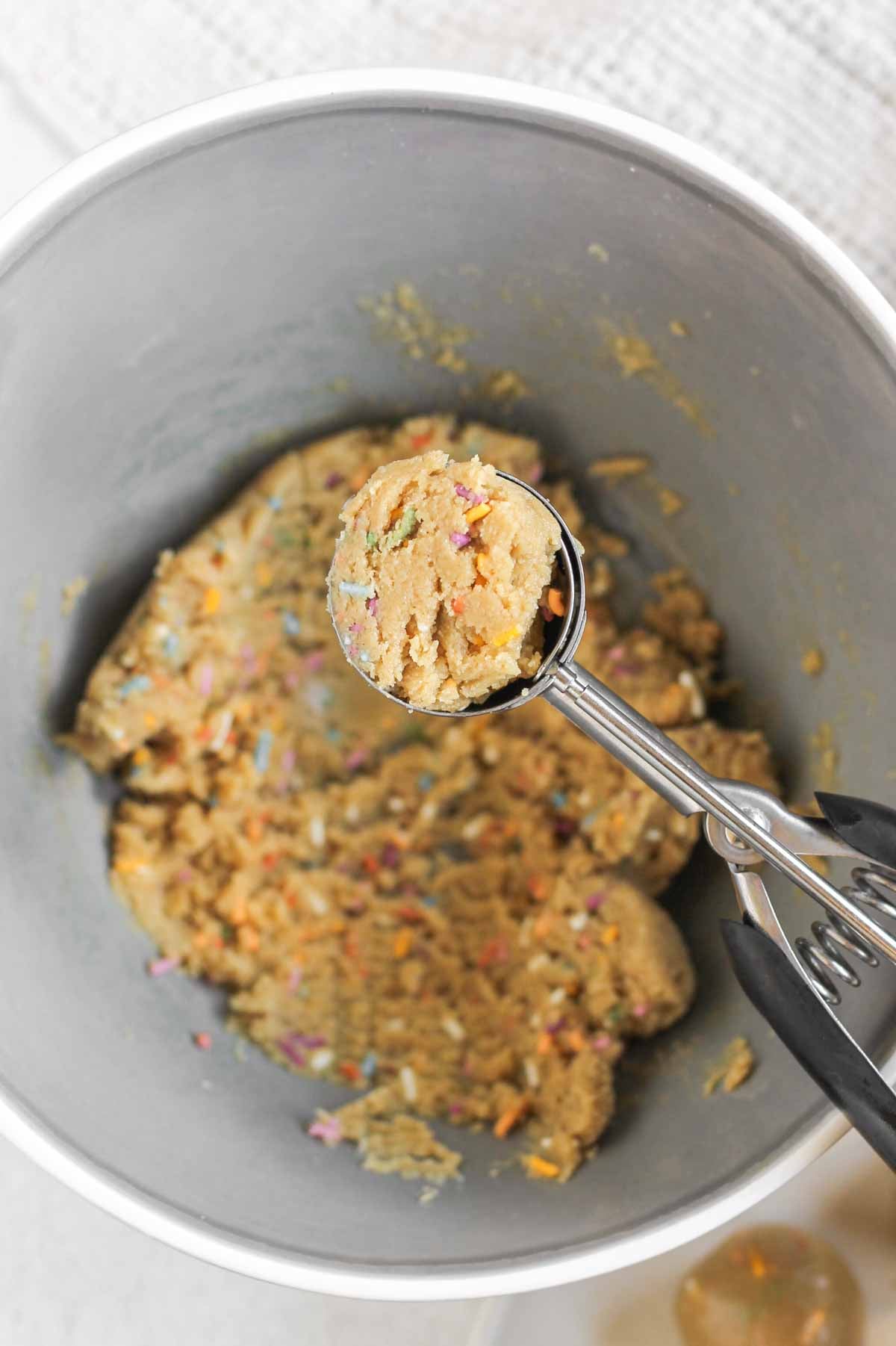 Spring-loaded scoop scooping out birthday cake protein ball mix from a mixing bowl.