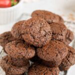 Mini chocolate muffins stacked on a wire cooling rack with a bowl of strawberries in the background.