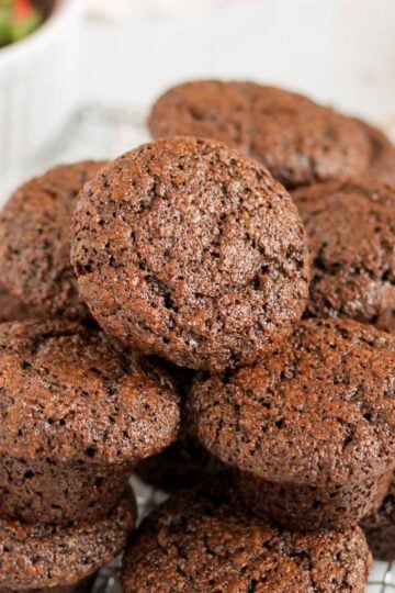 Mini chocolate muffins stacked on a wire cooling rack with a bowl of strawberries in the background.
