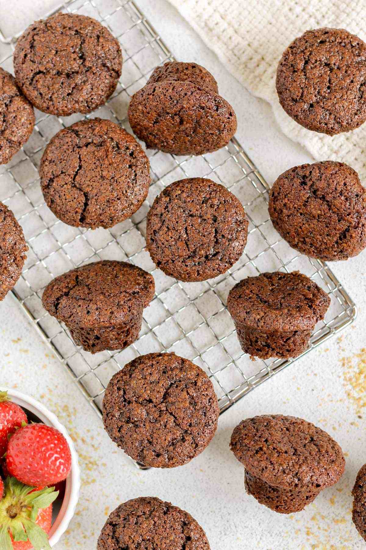 Mini chocolate flaxseed muffins spread out on a small wire cooling rack with a bowl of strawberries on the side.