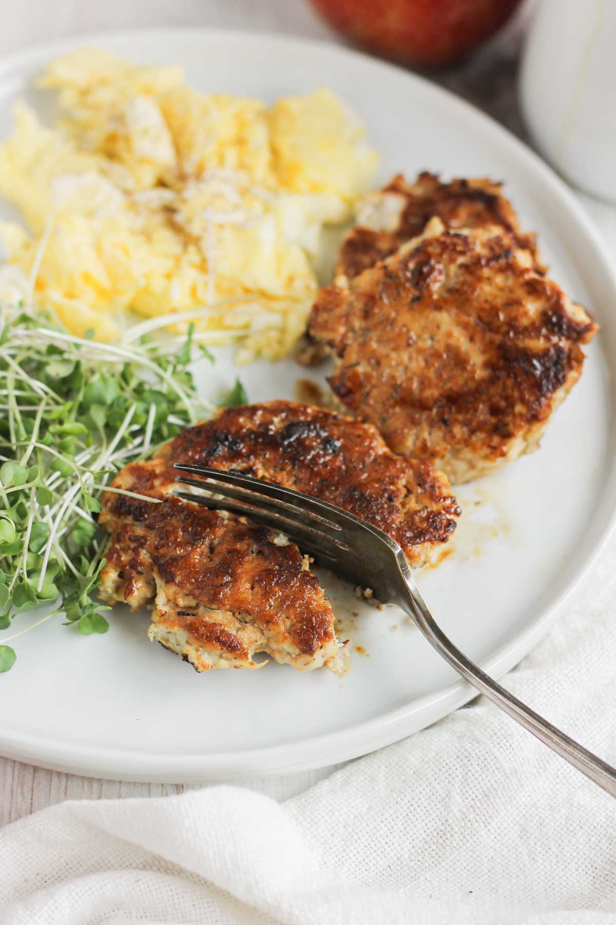 A fork cutting a chicken sausage patty on a plate with eggs and greens.