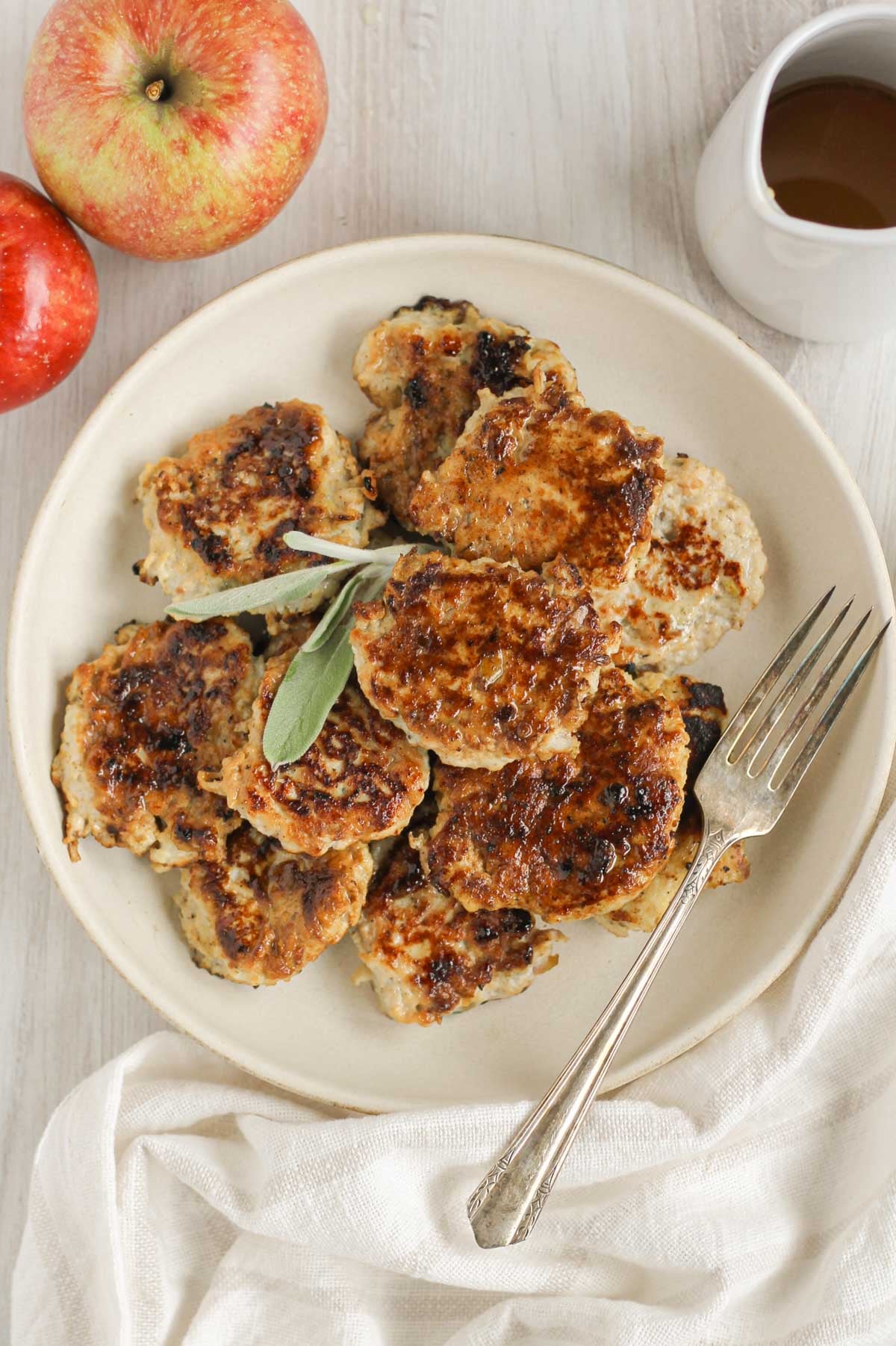 Chicken breakfast sausage patties on a round plate topped with a sprig of fresh sage with a fork on the plate and apples and maple syrup on the side.
