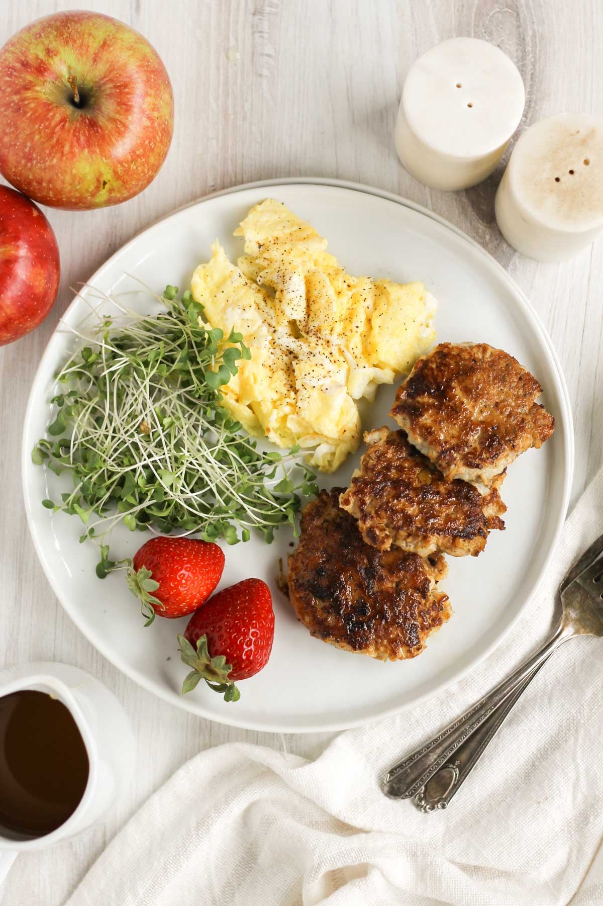 A plate with 3 chicken sausage patties, scrambled eggs, greens, and strawberries with apples and salt and pepper shakers on the side.