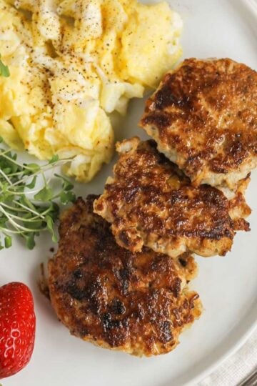 A plate with 3 chicken sausage patties, scrambled eggs, greens, and strawberries.
