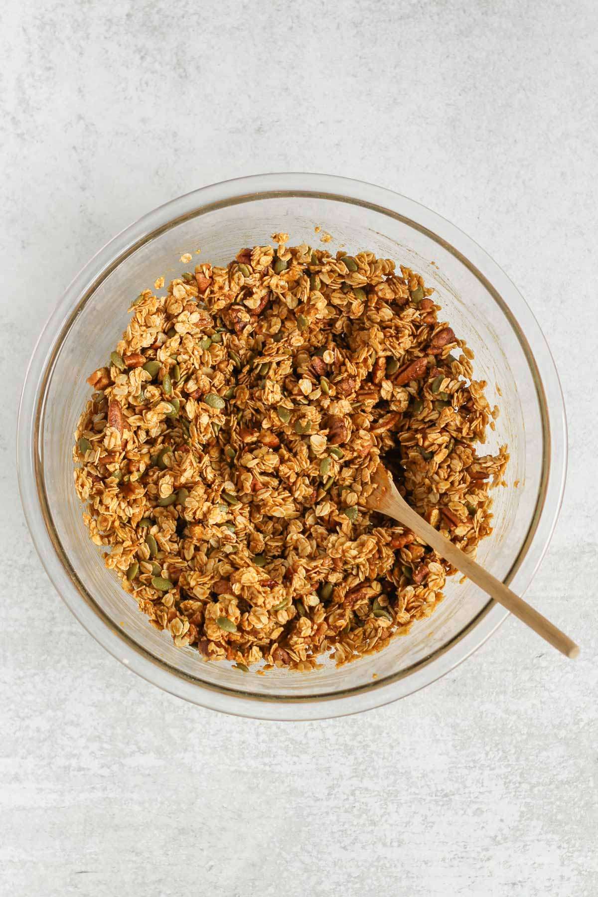 Pumpkin granola ingredients mixed together in a glass mixing bowl with a wooden spoon.