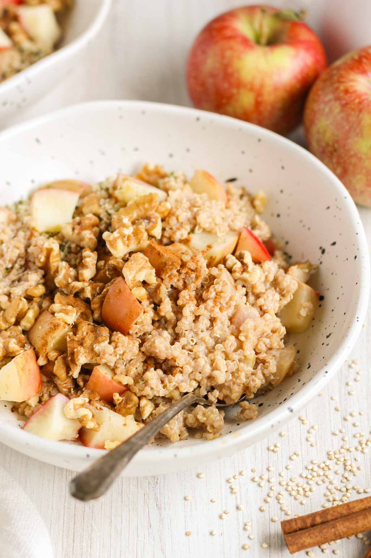 Apple cinnamon quinoa breakfast bowl topped with walnuts and sprinkled with cinnamon in a shallow bowl with apples in the background.