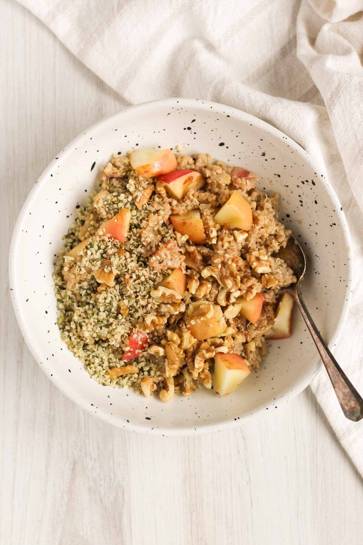 Cooked quinoa topped with cooked apples, walnuts, hempseeds and cinnamon in a shallow bowl with a spoon in the bowl.