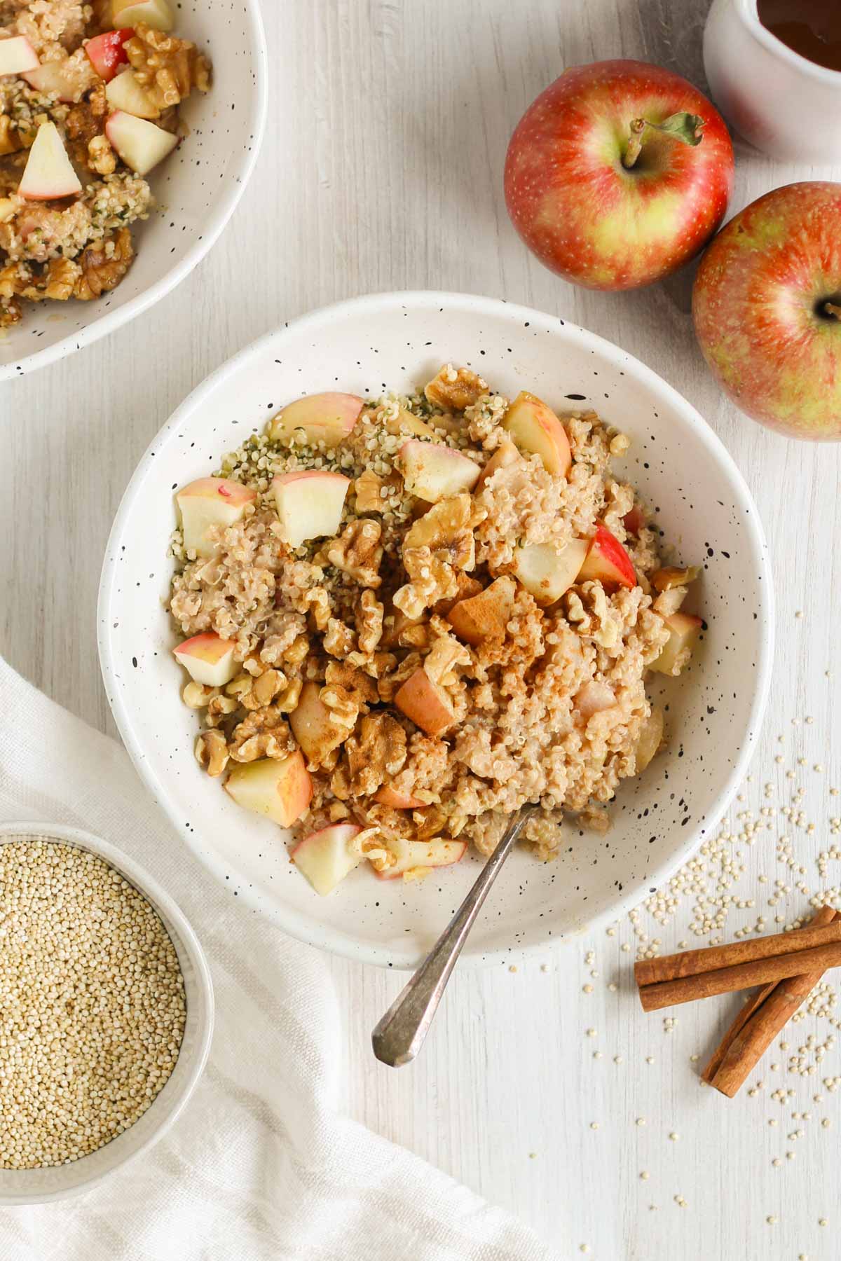 Apple cinnamon quinoa breakfast bowl topped with walnuts and sprinkled with cinnamon in a shallow bowl with a spoon in the bowl. Apples, quinoa, and cinnamon sticks off to the side.