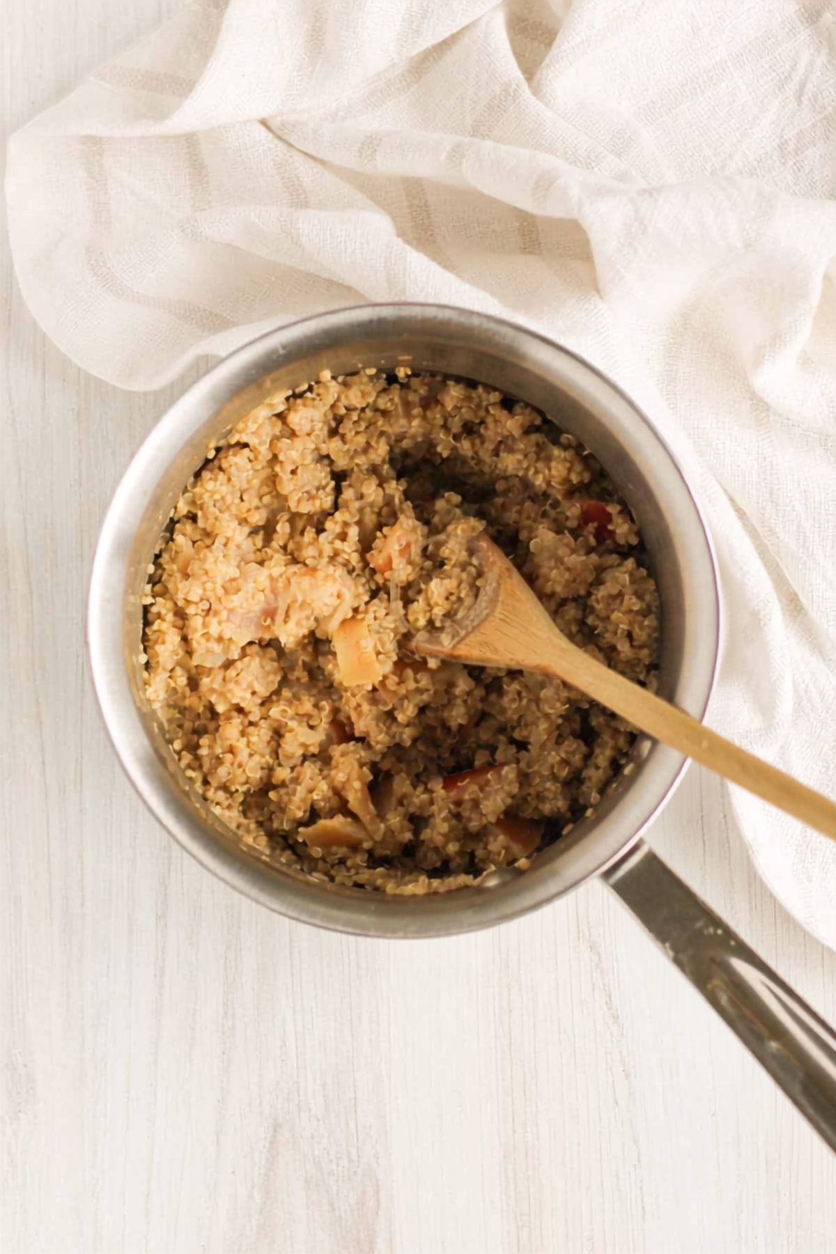 Cooked apple cinnamon quinoa in a stainless steel saucepan with a wooden spoon in the quinoa.