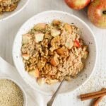 Apple cinnamon quinoa breakfast bowl topped with walnuts and sprinkled with cinnamon in a shallow bowl with a spoon in the bowl. Apples, quinoa, and cinnamon sticks off to the side.