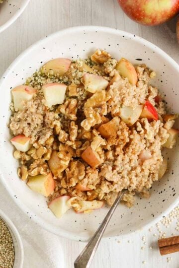 Apple cinnamon quinoa breakfast bowl topped with walnuts and sprinkled with cinnamon in a shallow bowl with a spoon in the bowl. Apples, quinoa, and cinnamon sticks off to the side.
