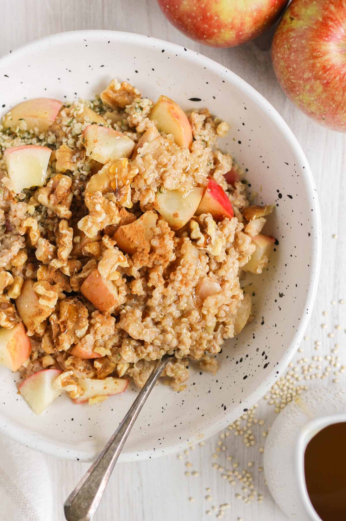 Apple cinnamon quinoa breakfast bowl topped with walnuts and sprinkled with cinnamon in a shallow bowl with a spoon in the bowl. Apples and a maple syrup on the side of the bowl.