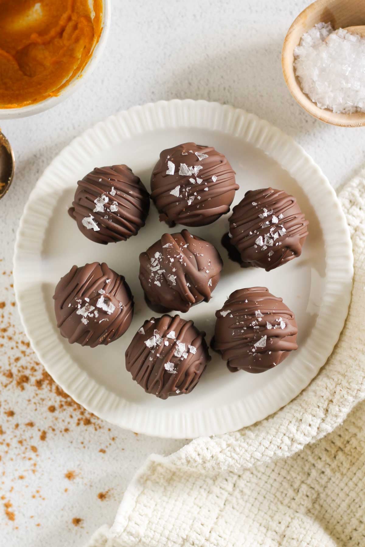 Seven pumpkin truffles topped with sea salt on a small round plate with a bowl of pumpkin and sea salt flakes off to the side.