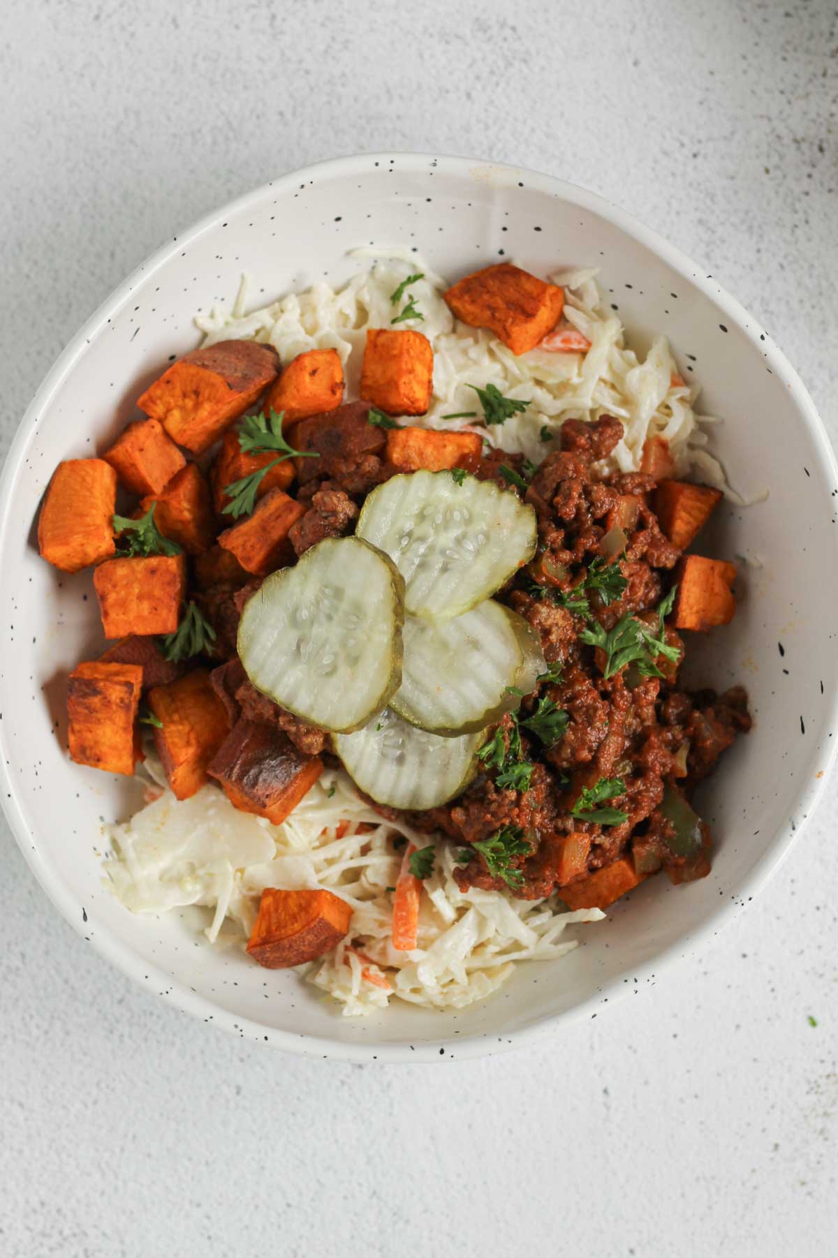 Shallow bowl with sloppy Joe mis, coleslaw, roasted sweet potatoes and pickles.