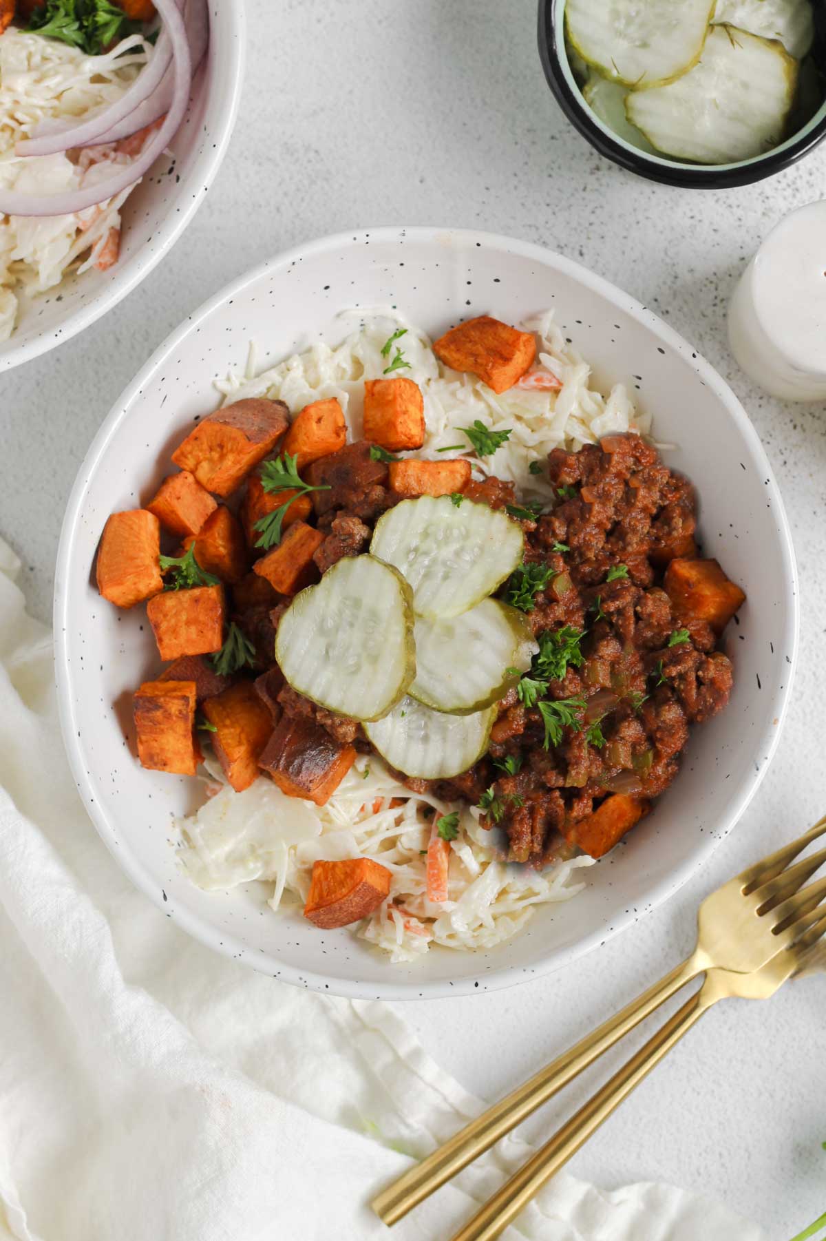 Shallow bowl with sloppy Joe mix, roasted sweet potatoes, coleslaw, and pickles with another bowl and a dish of pickles off to the side. Two gold forks laying next to the bowl.