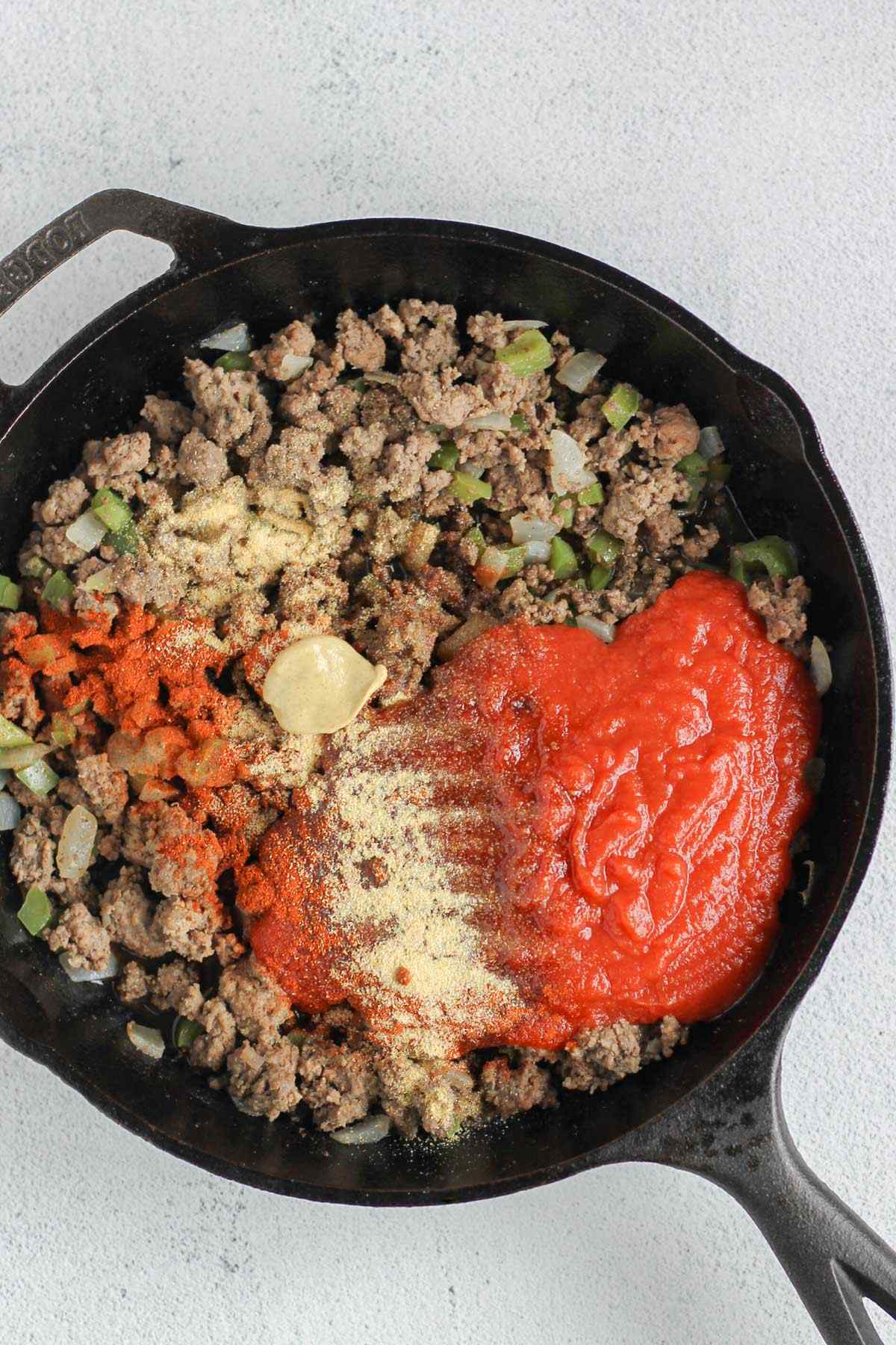 Cooked ground beef, onions, and peppers in a case iron skillet with sloppy Joe sauce ingredients added to the skillet.