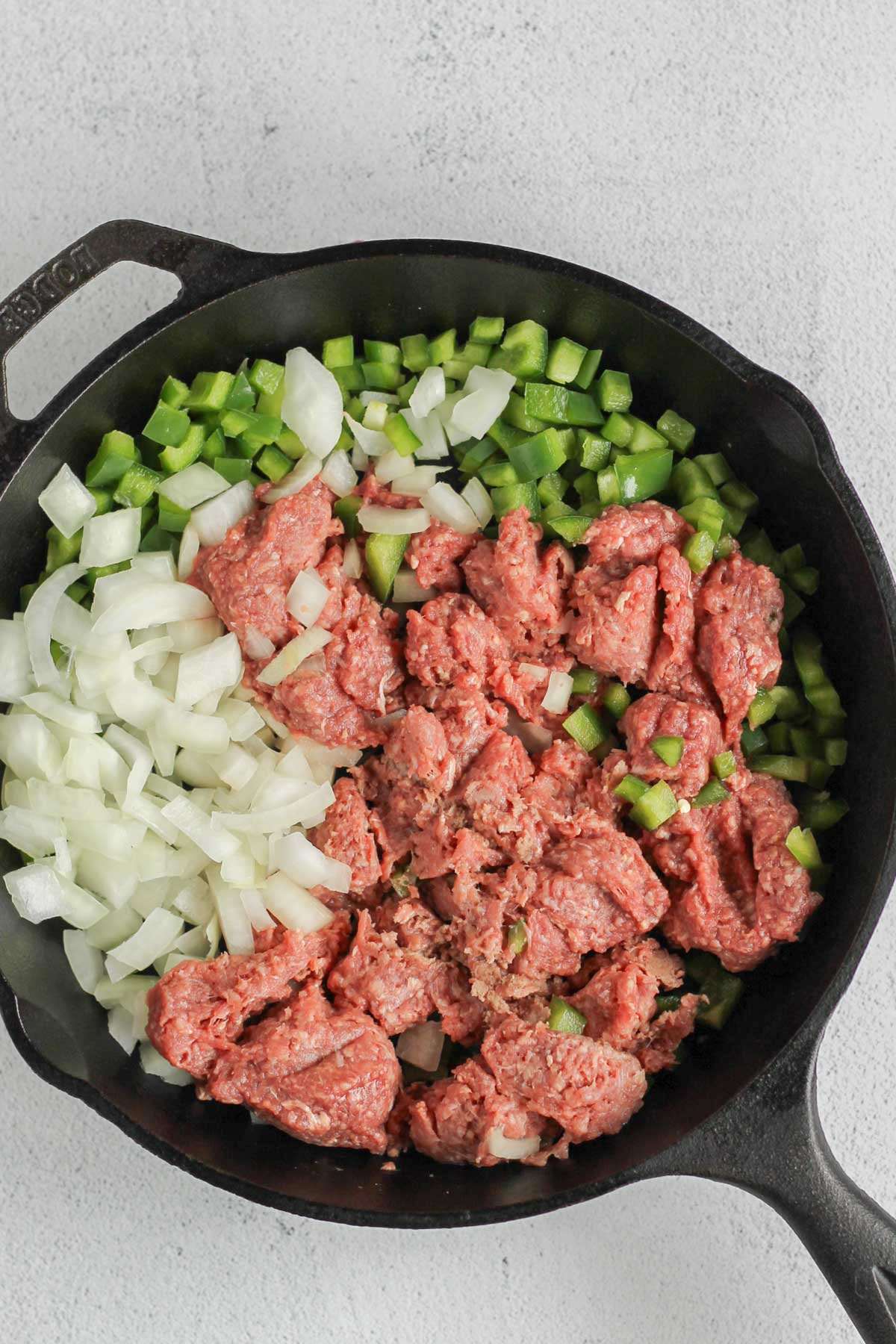 Raw ground beef, chopped onions, and chopped green peppers in a cast iron skillet.