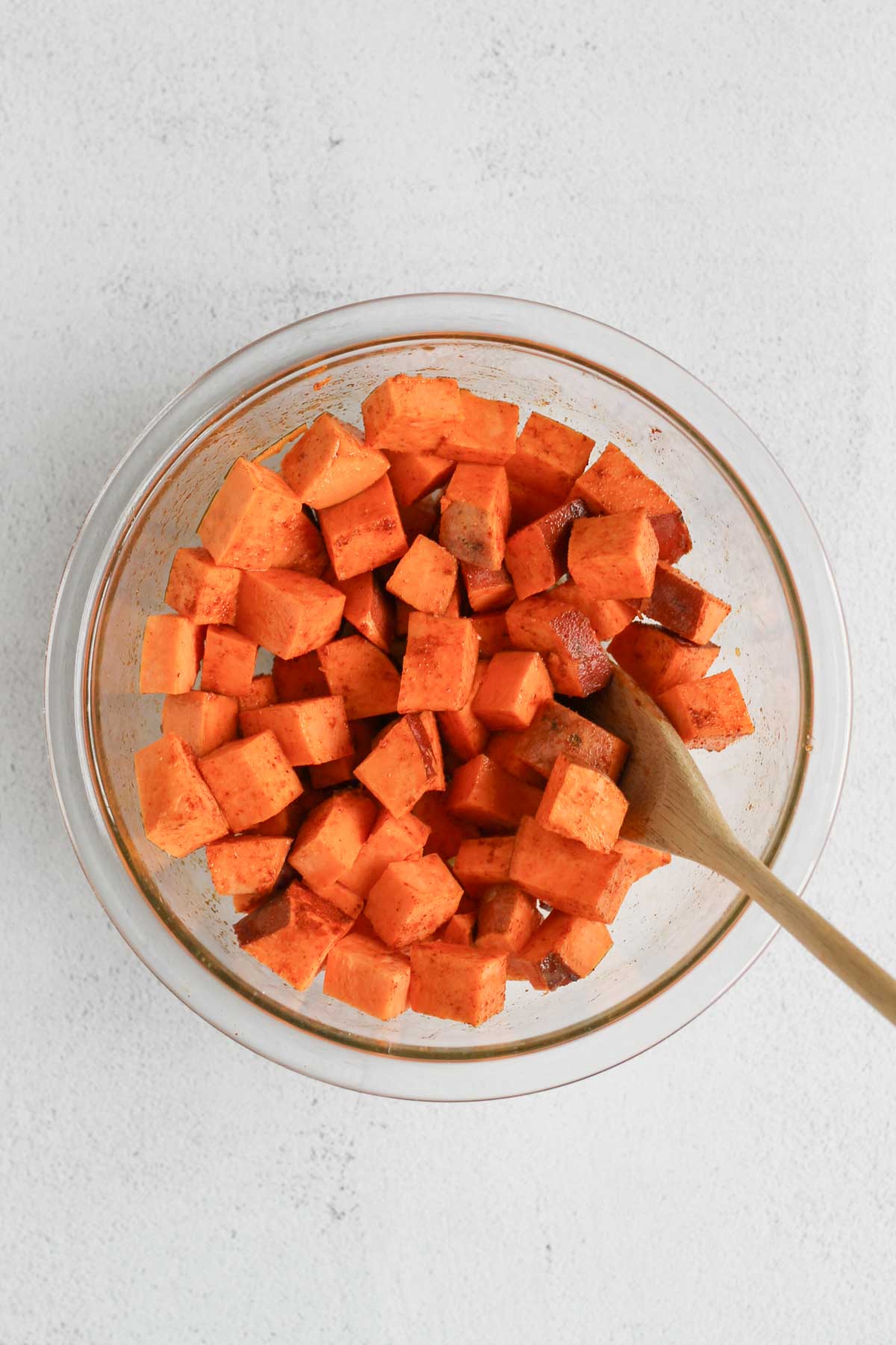 Chopped sweet potatoes mixed with olive oil and seasoning in a small glass mixing bowl with a wooden spoon in the bowl.