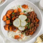 Shallow bowl with sloppy Joe mix, roasted sweet potatoes, coleslaw, and pickles with 2 gold forks on the side.